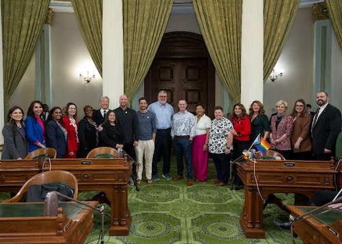 AD76 Patel Group photo of Assemblymembers including Dr. Darshana Patel third from left along with Teacher of the Year honorees on the Assembly floor. The Assembly floor has green carpet with gold draperies in the background and stately wooden desks in the foreground