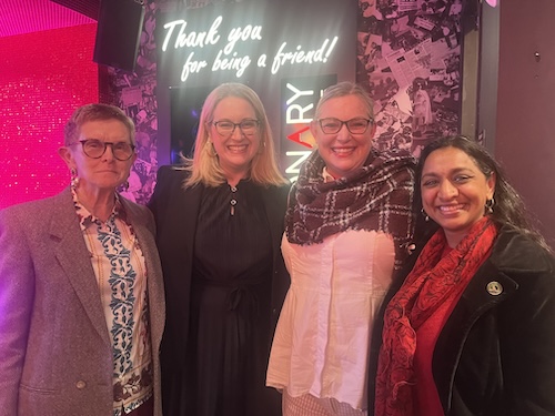 AD76 Patel Group photo of four women in a room with pink lighting and pink walls. Assemblymember Dr. Darshana Patel is far right wearing a black overcoat, red sweater, and red and black scarf. A white neon sign in the background reads Thank you for being a friend in handwritten-looking script.