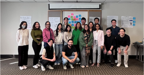 AD76 Patel Group photo of individuals of Asian descent in three rows with Assemblymember Dr. Darshana Patel second row, third from right wearing a pink jacket and grey pants. Post-it notes and large papers with writing on them are hung on the wall behind the people.