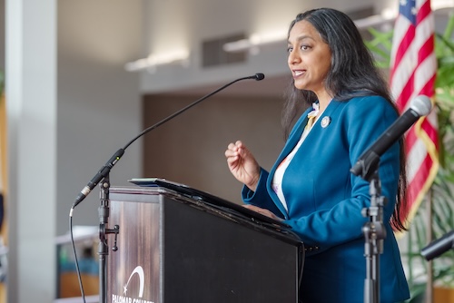 AD76 Patel Assemblymember Dr. Darshana Patel stands behind a podium and microphone delivers an address to attendees at the Community Swearing-In Ceremony. Assemblymember is photographed in ¾ profile from the waist up. American flag and tropical plants in background.