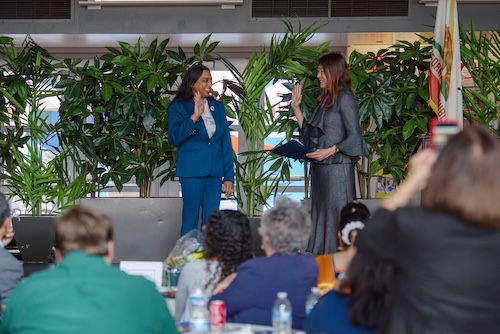 AD76 Patel Assemblymember Dr. Darshana Patel at left in three quarter profile wearing a teal suit with right hand raised, received the Oath of Office. She is smiling in front of a large audience on an elevated stage. State Treasurer Fiona Ma at right in three quarter profile wearing a grey suit administers the Oath of Office. Tropical plants and a California state flag in background. Audience members at tables in foreground.