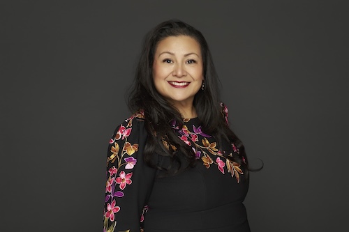 AD76 Patel Headshot of Dr. Star Rivera-Lacey smiling in three quarter profile with a black background and wearing a black dress with colorful embroidered flowers.